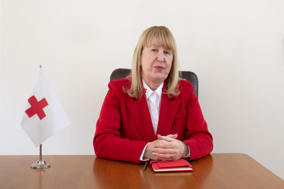 Tanya Petrova Georgieva is sitting at a desk next to a small red cross flag and wearing a red vest.