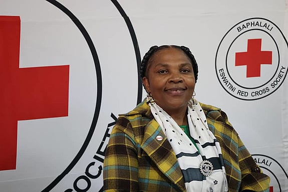 Happy Nozizwe Dlamini, from the Eswatini Red Cross Society, looks into the camera and smiles. She is inside of Red Cross facilities and there is a big Red Cross in the background.